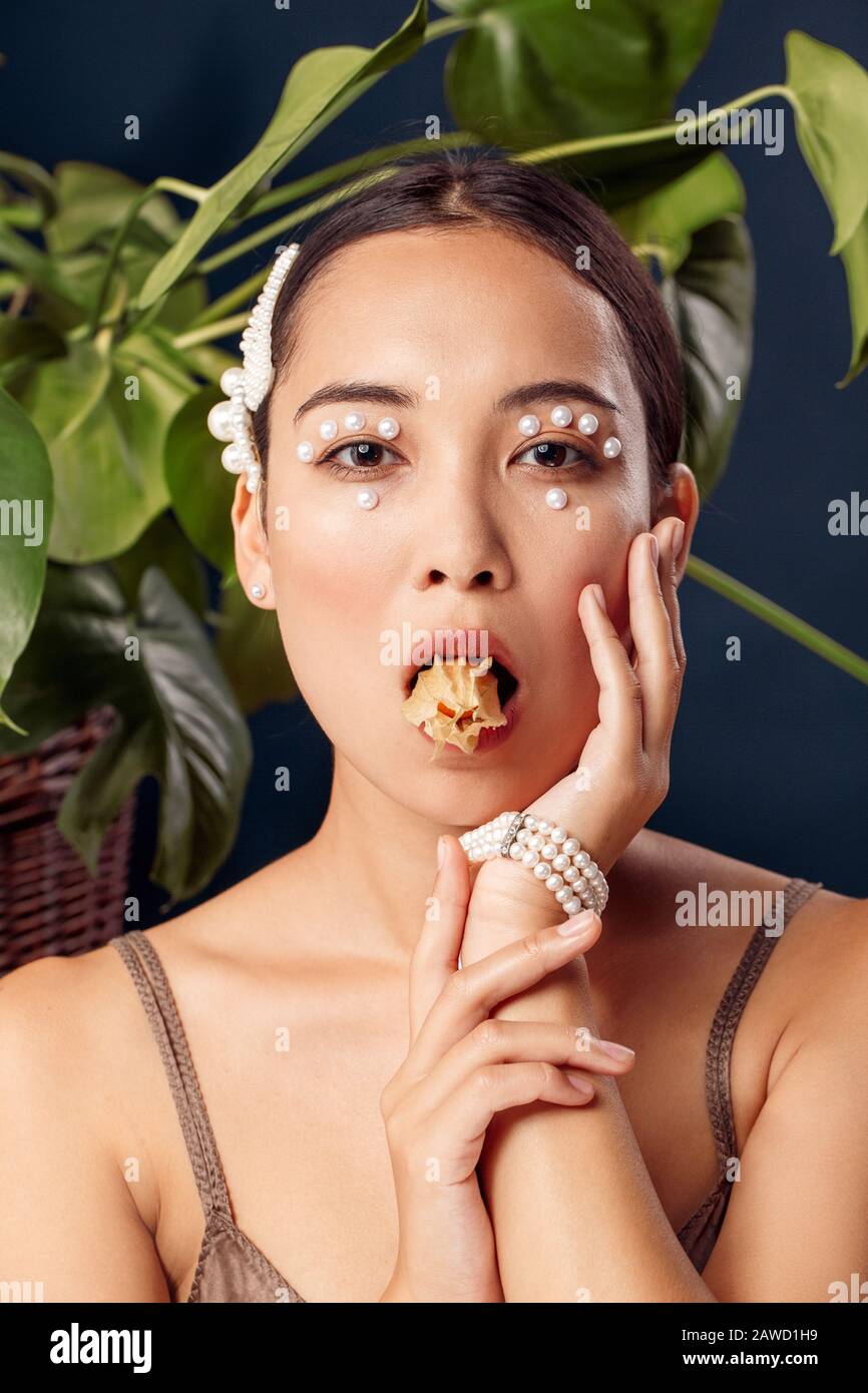 Freestyle. Young woman eyes decorated with perls wearing bracelet standing isolated on dark blue with plants holding physalis in mouth posing to Stock Photo