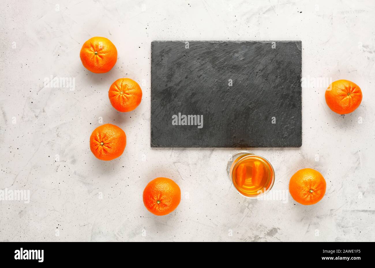 A cutting board made of gray slate is surrounded by orange fruits of mandarins randomly lying nearby on a gray concrete background, copy space. Stock Photo