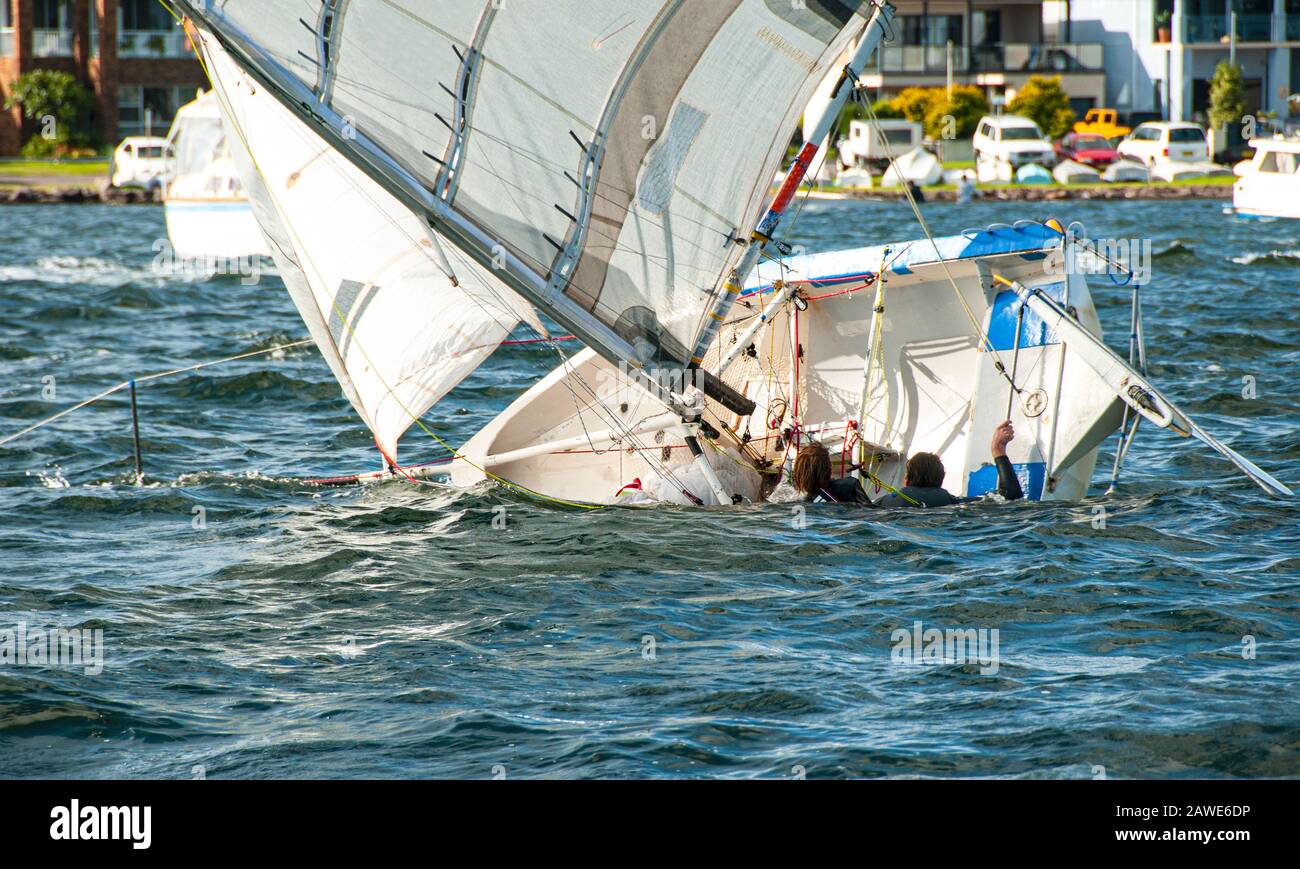 A girl and boy in the water climbing back into a capsized racing sailboat. Teamwork by junior sailors racing on saltwater Lake Macquarie. Stock Photo