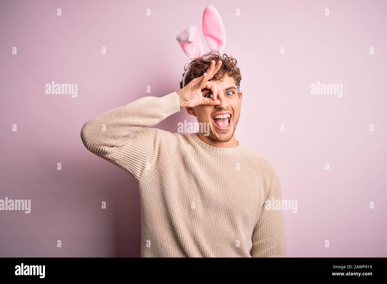 Young handsome blond man wearing easter rabbit ears over isolated pink background doing ok gesture with hand smiling, eye looking through fingers with Stock Photo