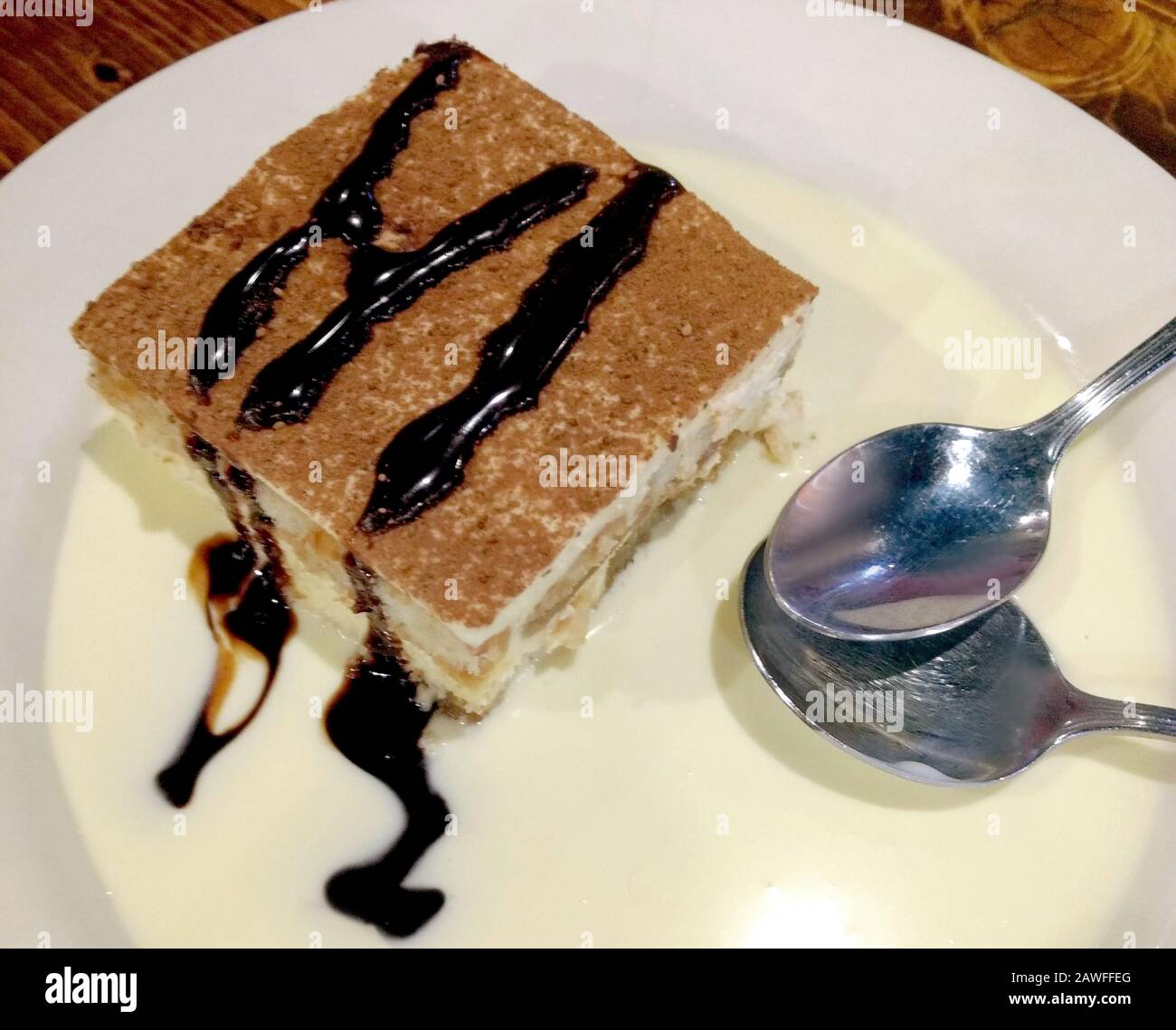 Delicious slice of tiramisu on a vanilla cream sauce, drizzled with chocolate and served with two spoons for sharing Stock Photo