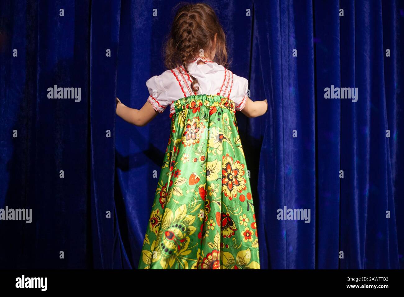 The child is waiting for a performance on stage. A girl peeks behind the curtain. Theatrical stage with a blue velvet curtain. Costume embroidered in Stock Photo