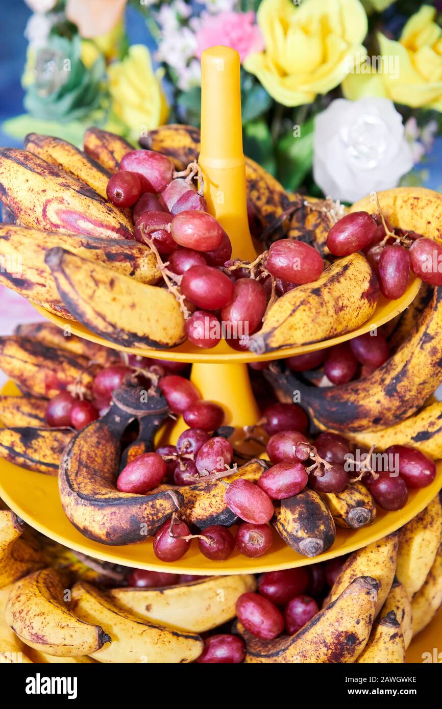 Close-up view of overripe spotted bananas and red grapes placed on a plastic tower tray, with colorful plastic flowers seen in the background Stock Photo