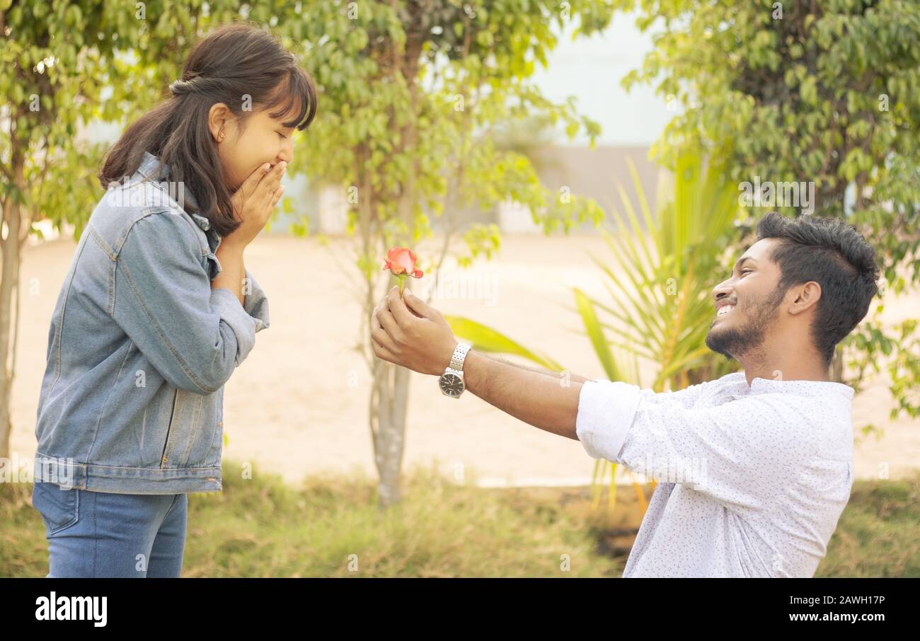 Concept of teenage love or affection - young man proposing to smiling excited girlfriend standing on knee with red rose on valentines day Stock Photo