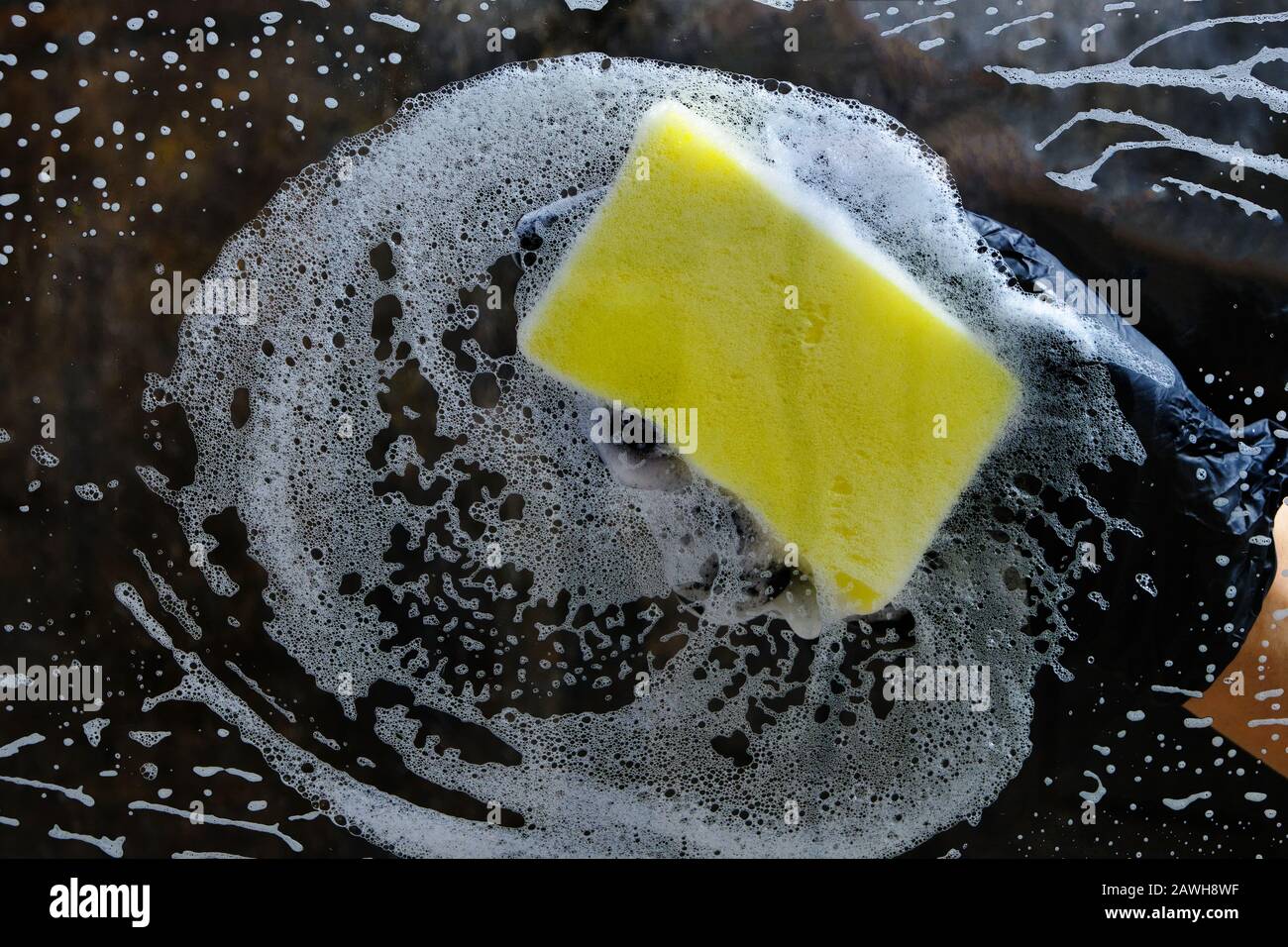 Hand in a black glove washes the glass with a foam sponge with foam on a dark background. Stock Photo
