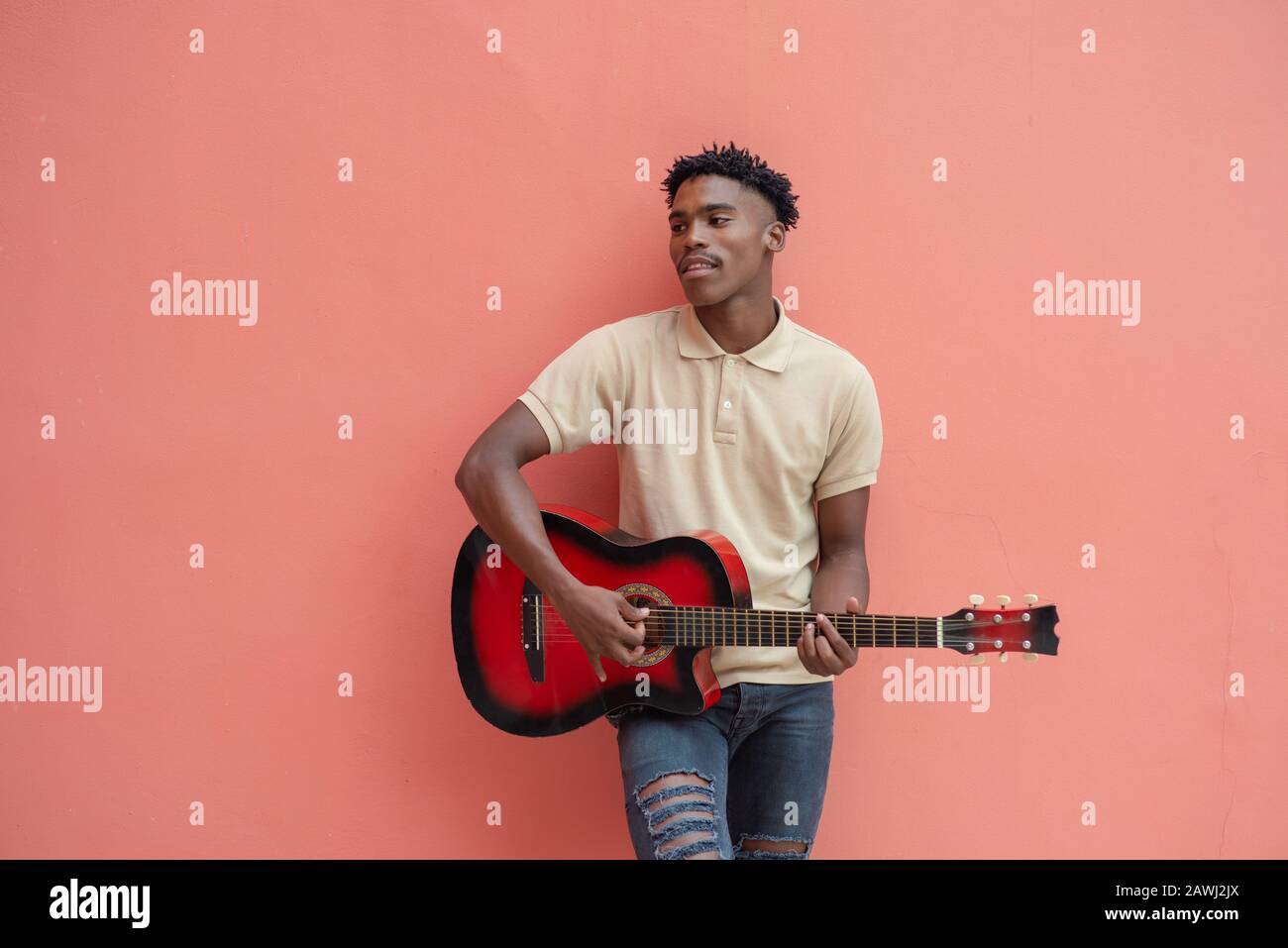 Young man with guitar standing against bright colored wall while performing Stock Photo