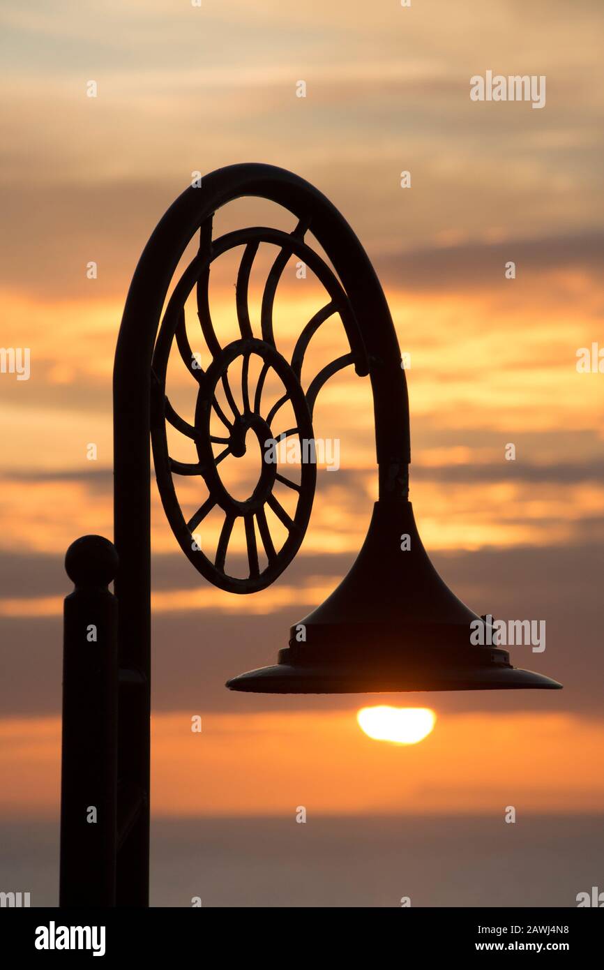 A lampost at sunrise with ammonite designs along the seafront of the town of Lyme Regis, the rising sun is just below the lamp. The town is situated o Stock Photo