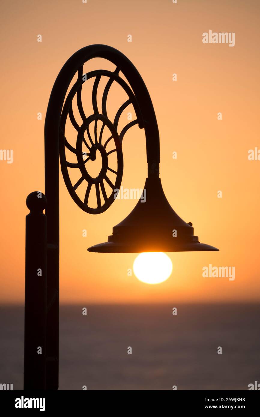 A lampost at sunrise with ammonite designs along the seafront of the town of Lyme Regis, the rising sun is just below the lamp. The town is situated o Stock Photo