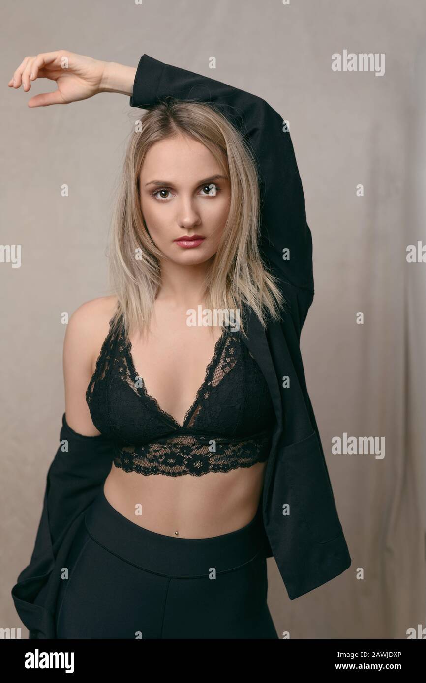 Sexy young blond woman in black lingerie stretching her arms above her head while looking at the camera with a serious sultry expression over a brown Stock Photo