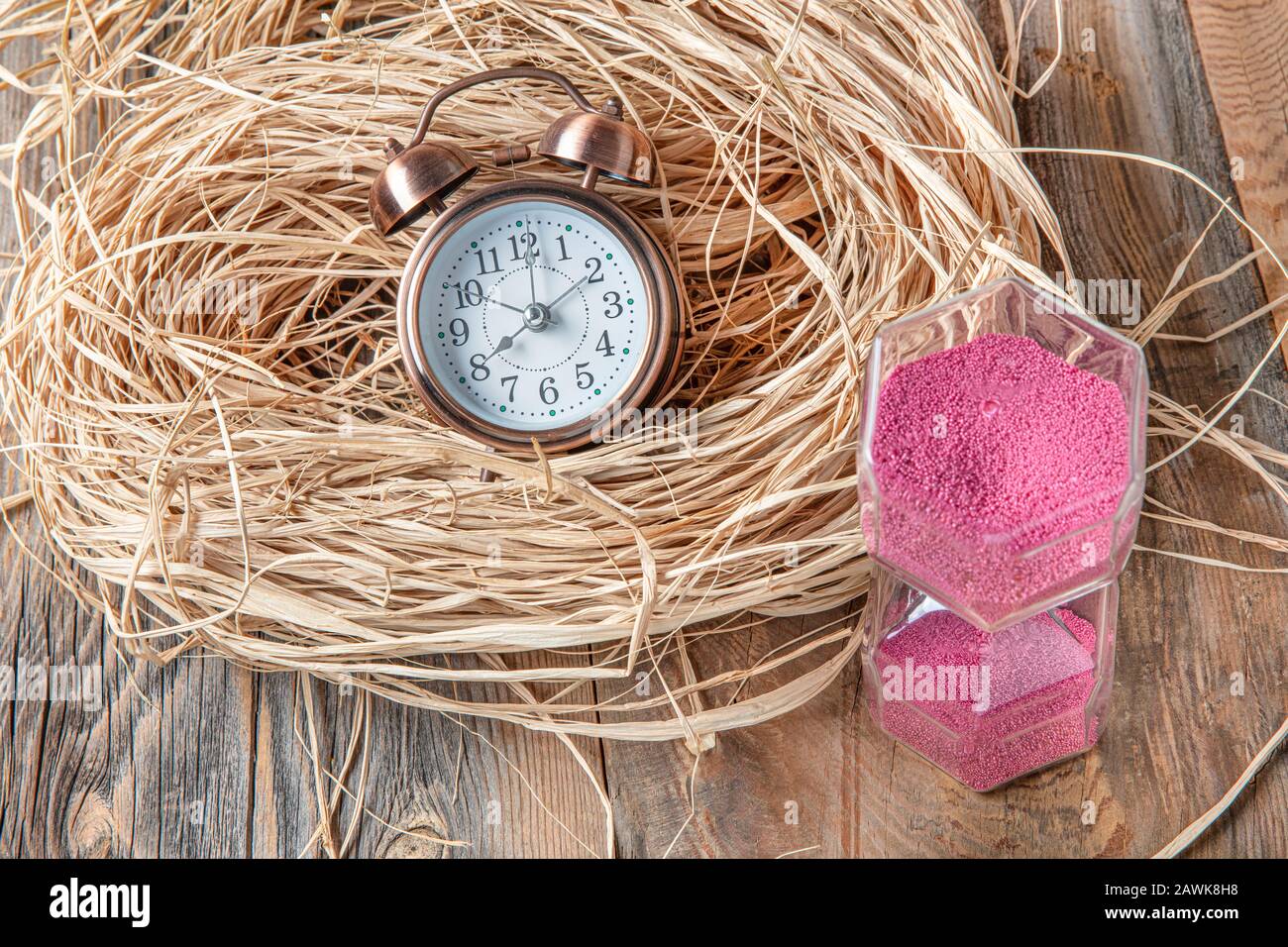 Pink hourglass and alarm clock on straw on wooden floor. Concept of