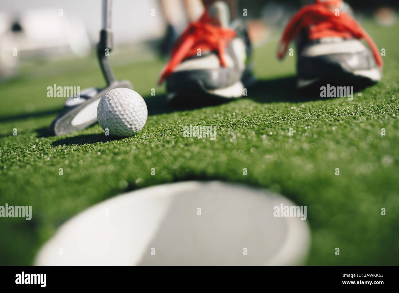Kids play mini golf. Close-up image of player in snickers with mini golf club and white golf ball. e cup in the foregroundGold hole th Stock Photo