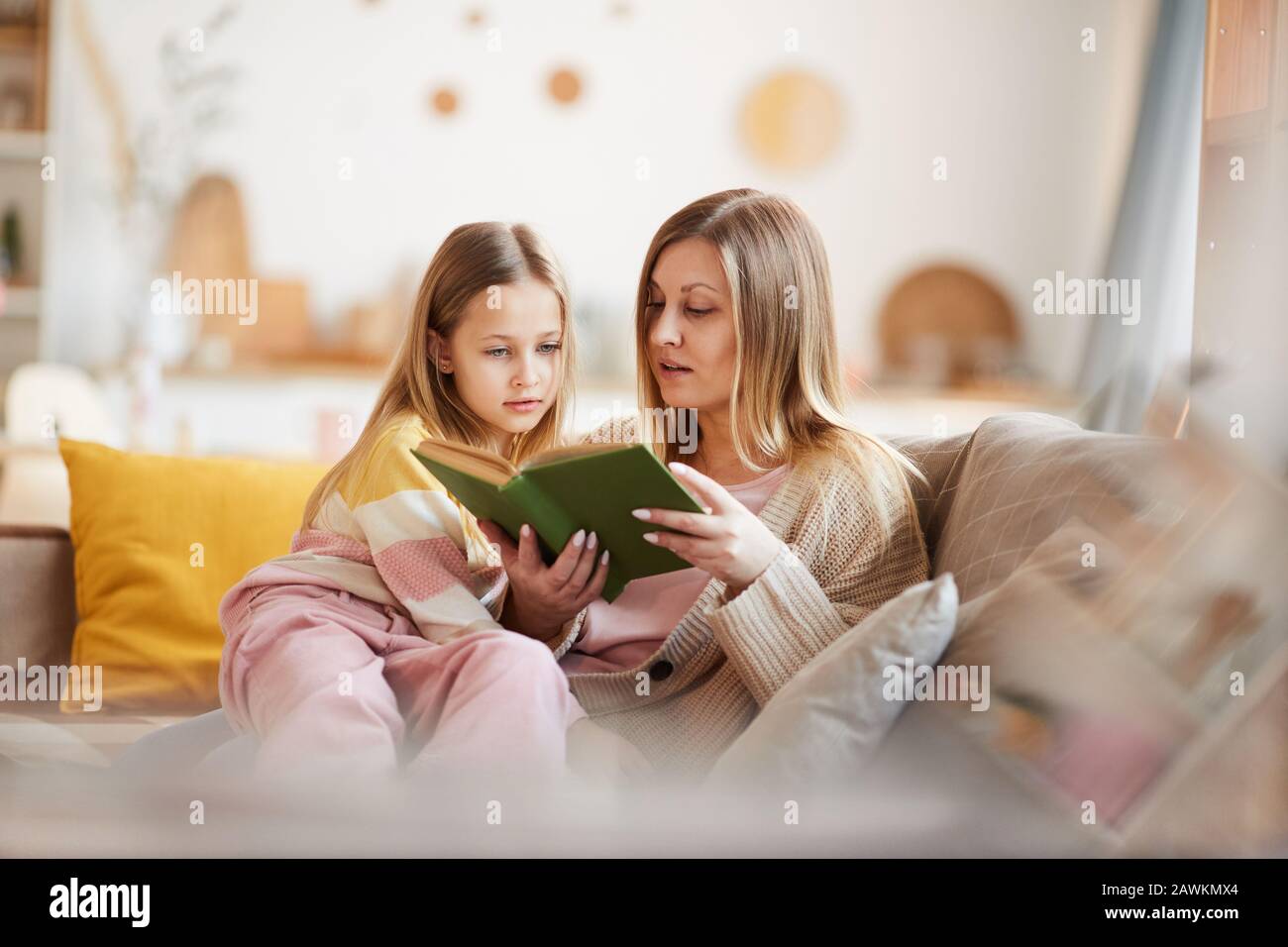 Warm-toned portrait of mature mother reading book to little girl while sitting on sofa in cozy home interior, copy space Stock Photo