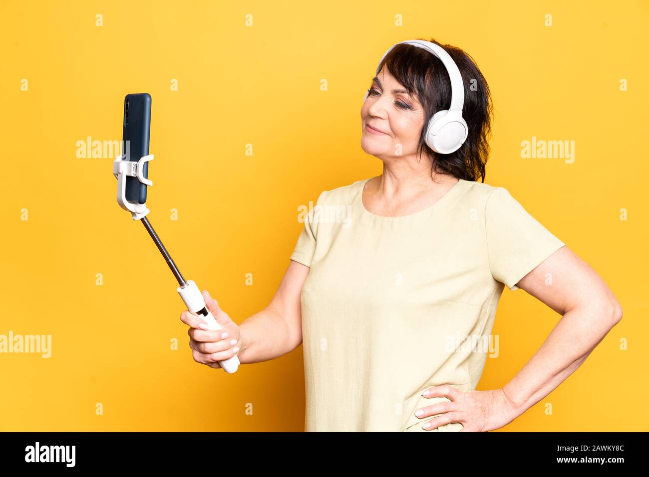 Beautiful Senior woman making video call by smartphone, monopod and wireless headphones over yellow background Stock Photo