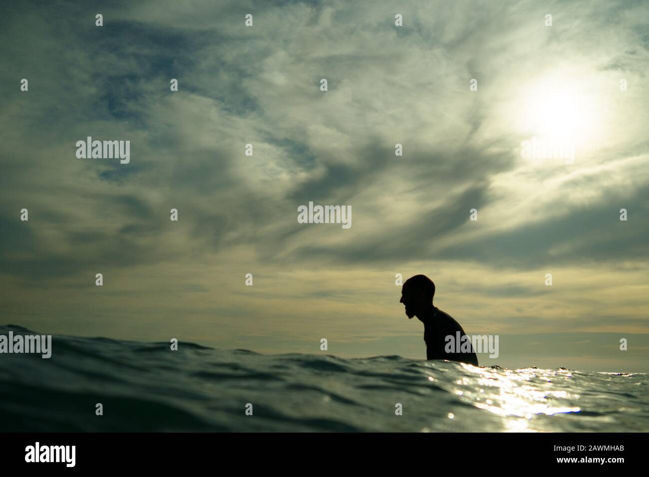 A young male surfer in a black wetsuit with a goatee beard sits in silhouette on a surfboard waiting for a wave at Piha Beach, Piha, West Auckland. Stock Photo