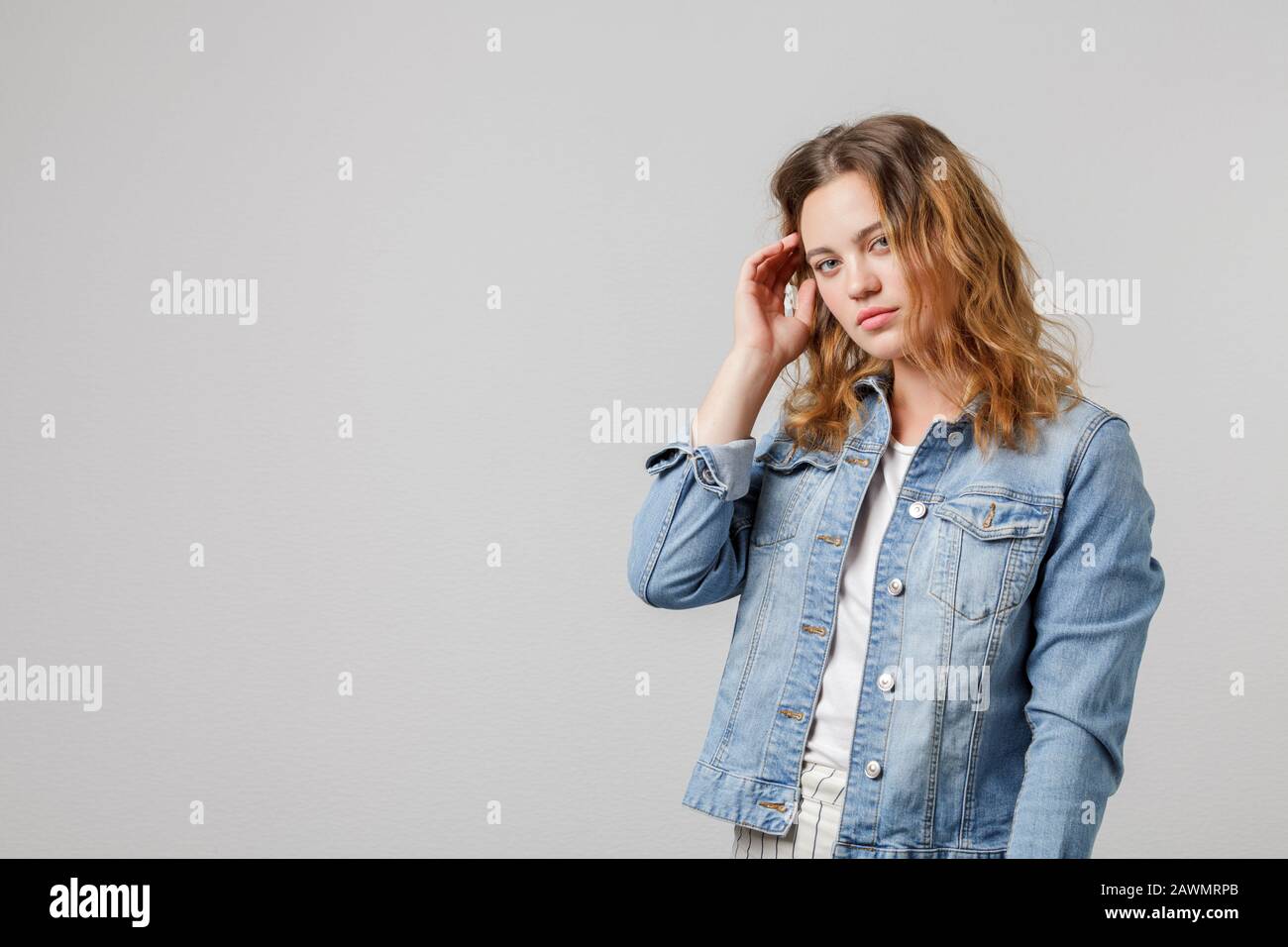 Young girl with long flowing hair in a denim jacket looks thoughtfully at the camera and touches her hair. Half-length portrait on a white background. Stock Photo