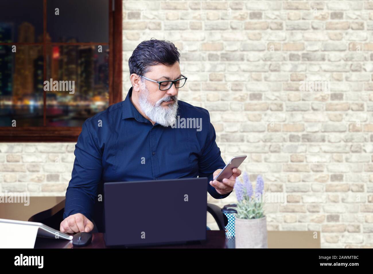 Successful mature middle eastern businessman wearing eyeglasses working on laptop while using phone in modern office Stock Photo