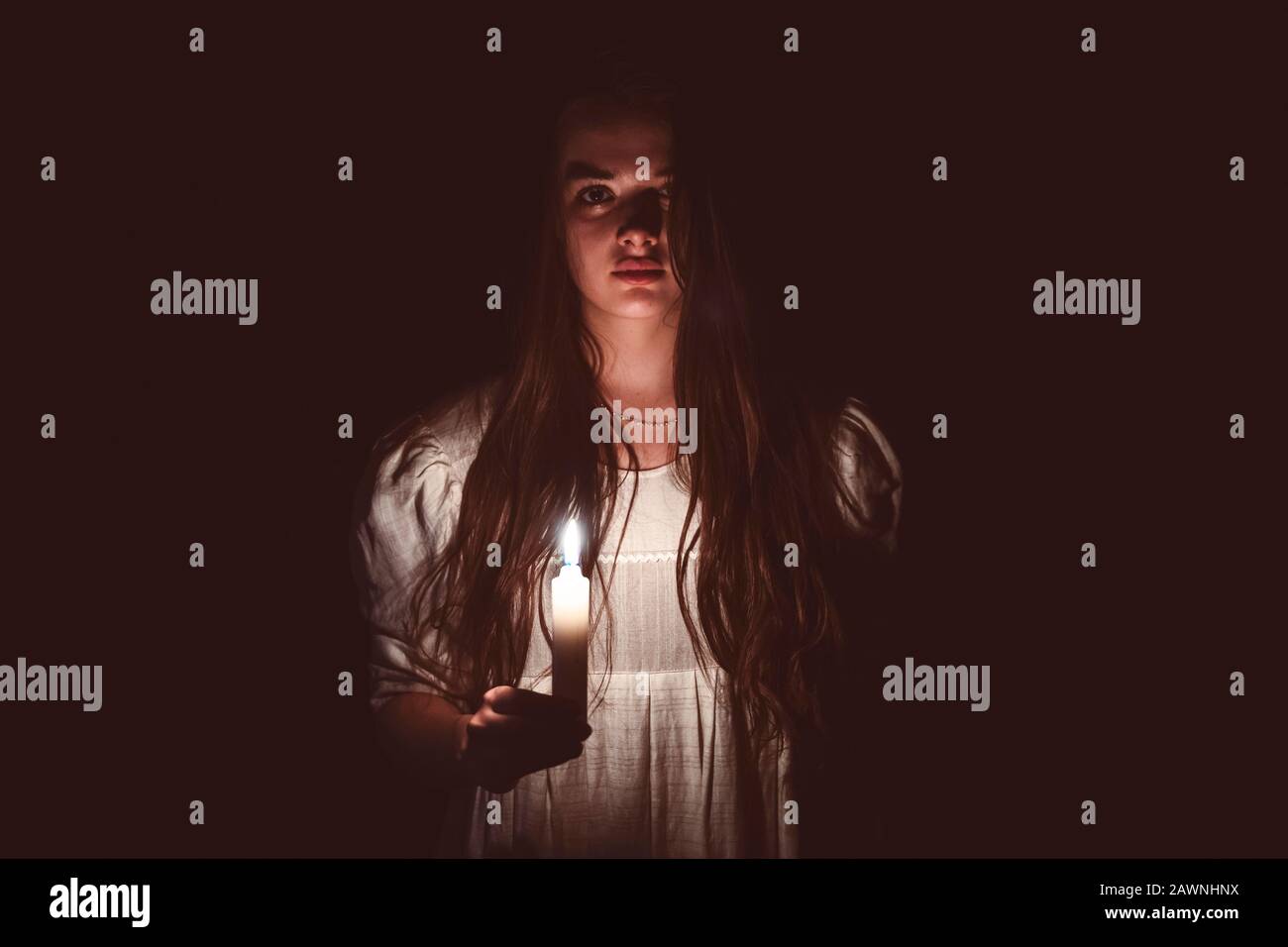 A young scary girl in an old white dress holding a candle in her hand and staring in to the camera. Dark background. Scary horror concept. Stock Photo