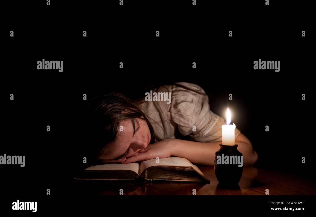 A young girl sleeping on an old book at night with candle light. The girl is wearing an old white dress. Dark background. Front view. Stock Photo