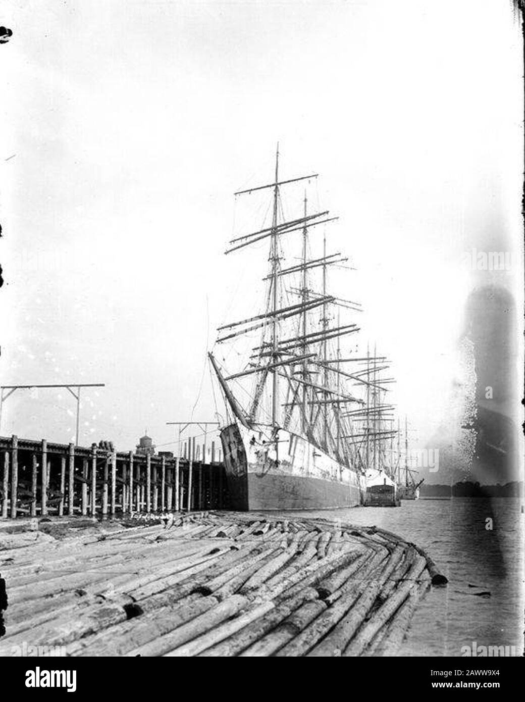 Four-masted square-rigger tied up at pier with log boom in foreground ...