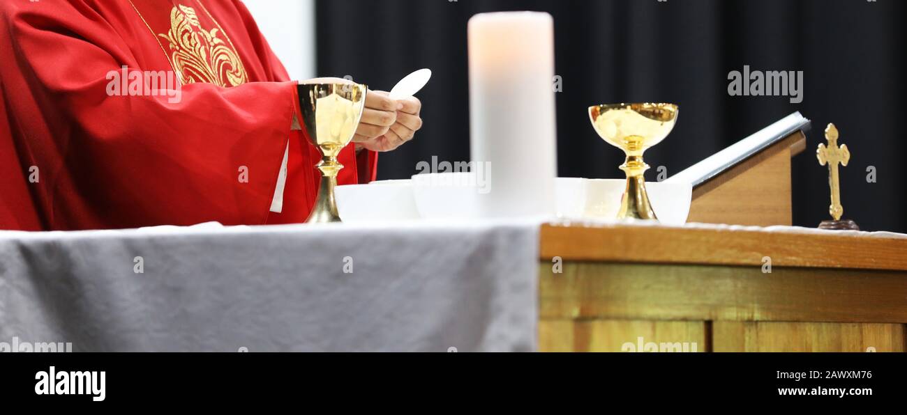 A Catholic Priest about to break the bread host while celebrating blessed Holy Communion at Mass. Wearing a red gown vestment and surrounded by chalic Stock Photo