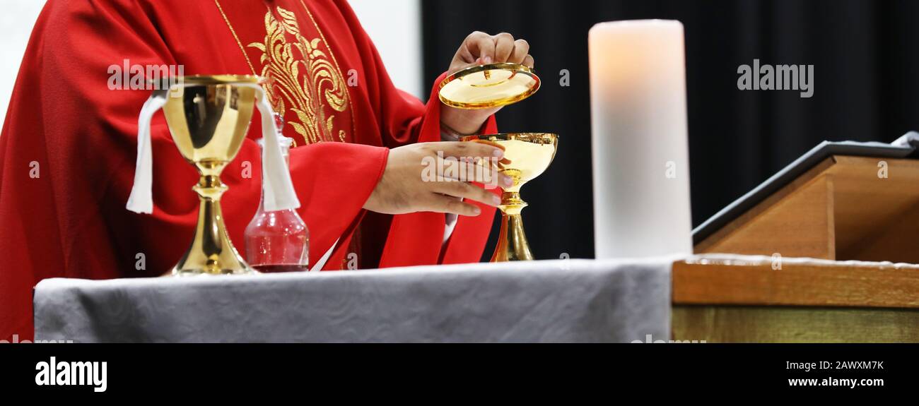 A Catholic Priest opening brass vessel containing bread host while celebrating blessed Holy Communion at Mass. Wearing a red gown vestment and surroun Stock Photo