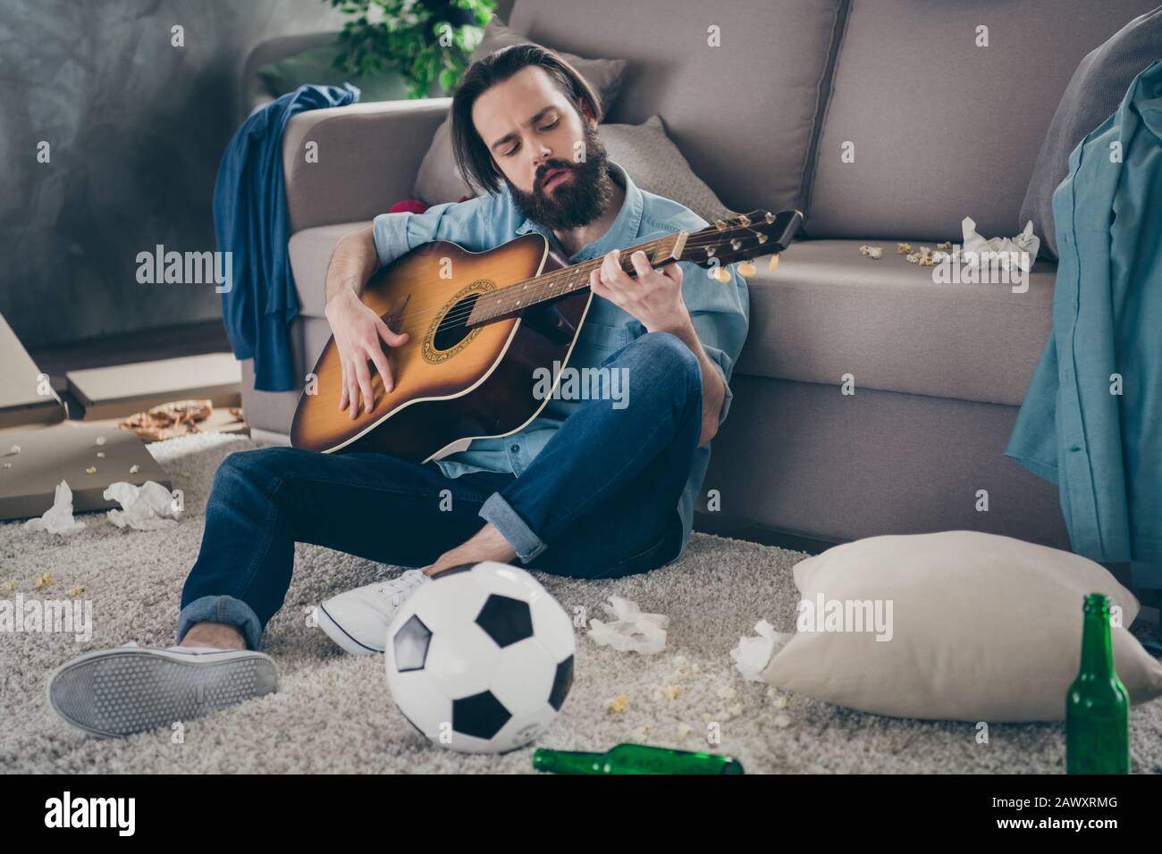 Photo of hipster guy with long beard sitting carpet near sofa holding guitar don't mind chaos after stag party messy dirty flat singing songs careless Stock Photo