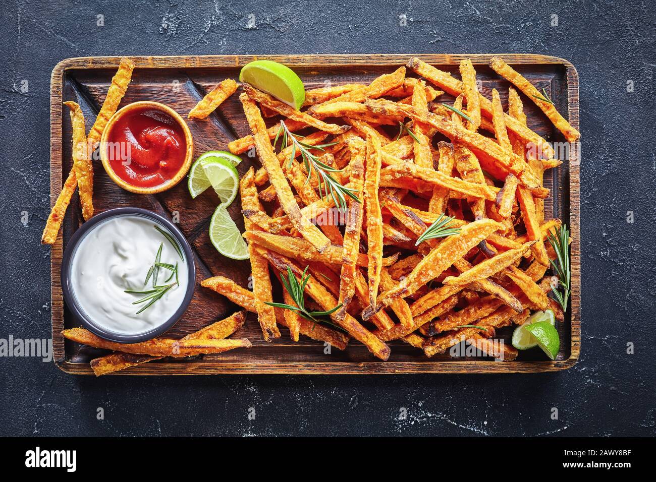 Sweet Potato Fries with sauce and ketchup on a rude wooden board on a concrete table with lime and bouquet of aromatic herbs, horizontal view from abo Stock Photo