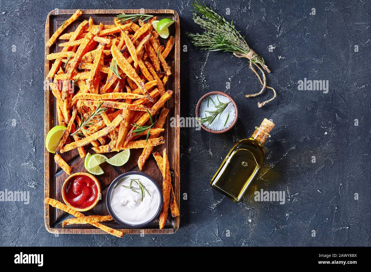 close-up of crispy salty Sweet Potato Fries with sauce and ketchup on a rude wooden board on a concrete table with lime and bouquet of aromatic herbs, Stock Photo