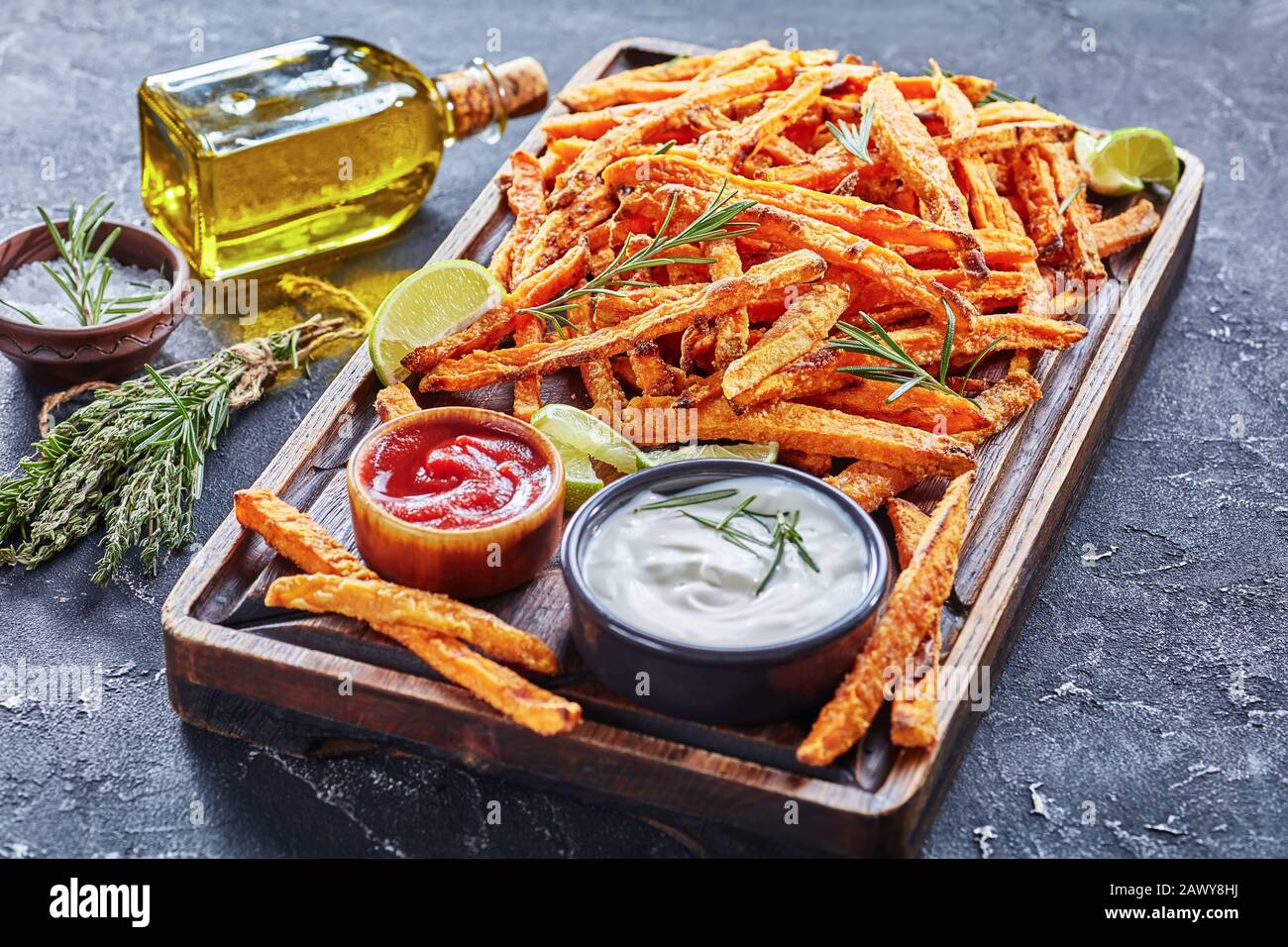 crispy salty Sweet Potato Fries with sauce and ketchup on a rude wooden board on a concrete table with lime and bouquet of aromatic herbs, horizontal Stock Photo