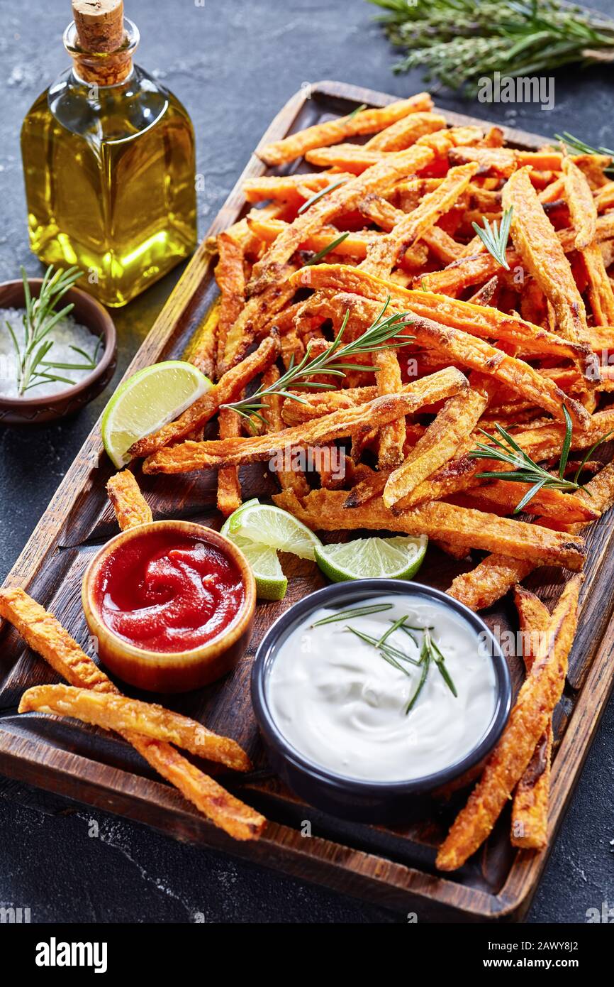 crispy salty Sweet Potato Fries with sauce and ketchup on a rude wooden board on a concrete table with lime and bouquet of aromatic herbs, vertical vi Stock Photo