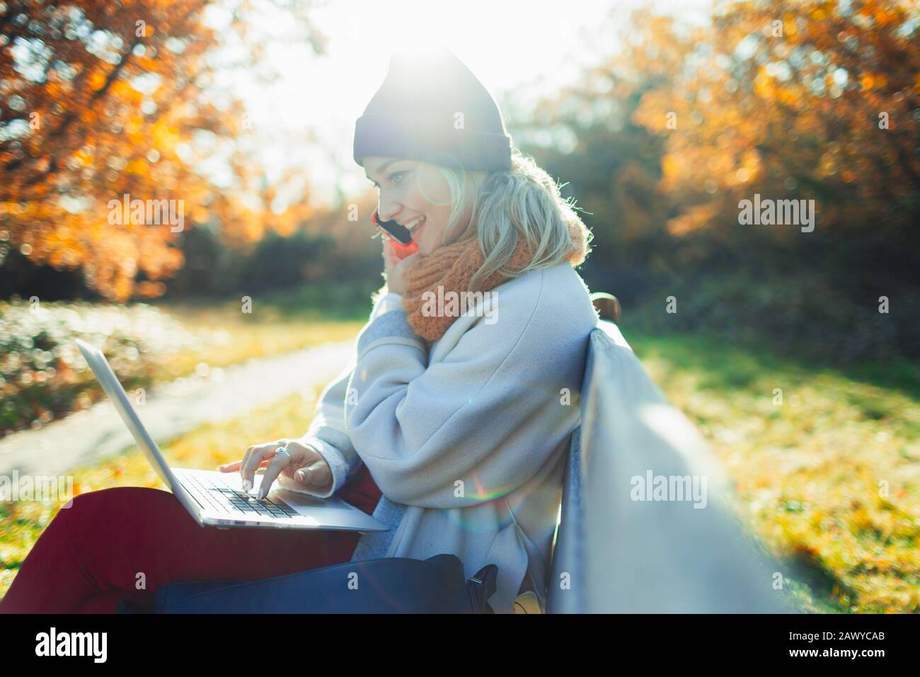 Young woman using laptop and talking on smart phone on sunny autumn park bench Stock Photo
