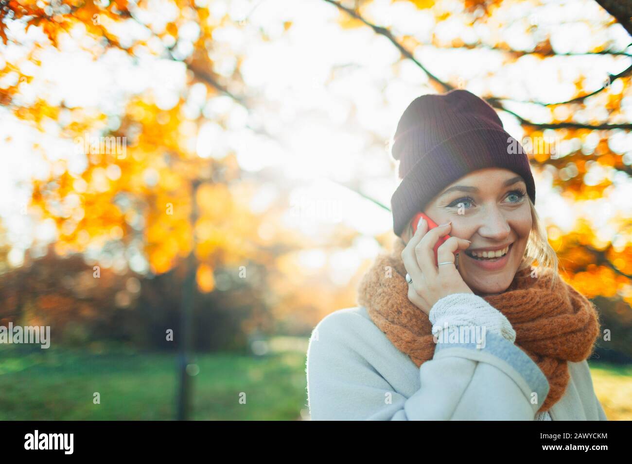 Young woman in stocking cap and scarf talking on smart phone in sunny autumn park Stock Photo