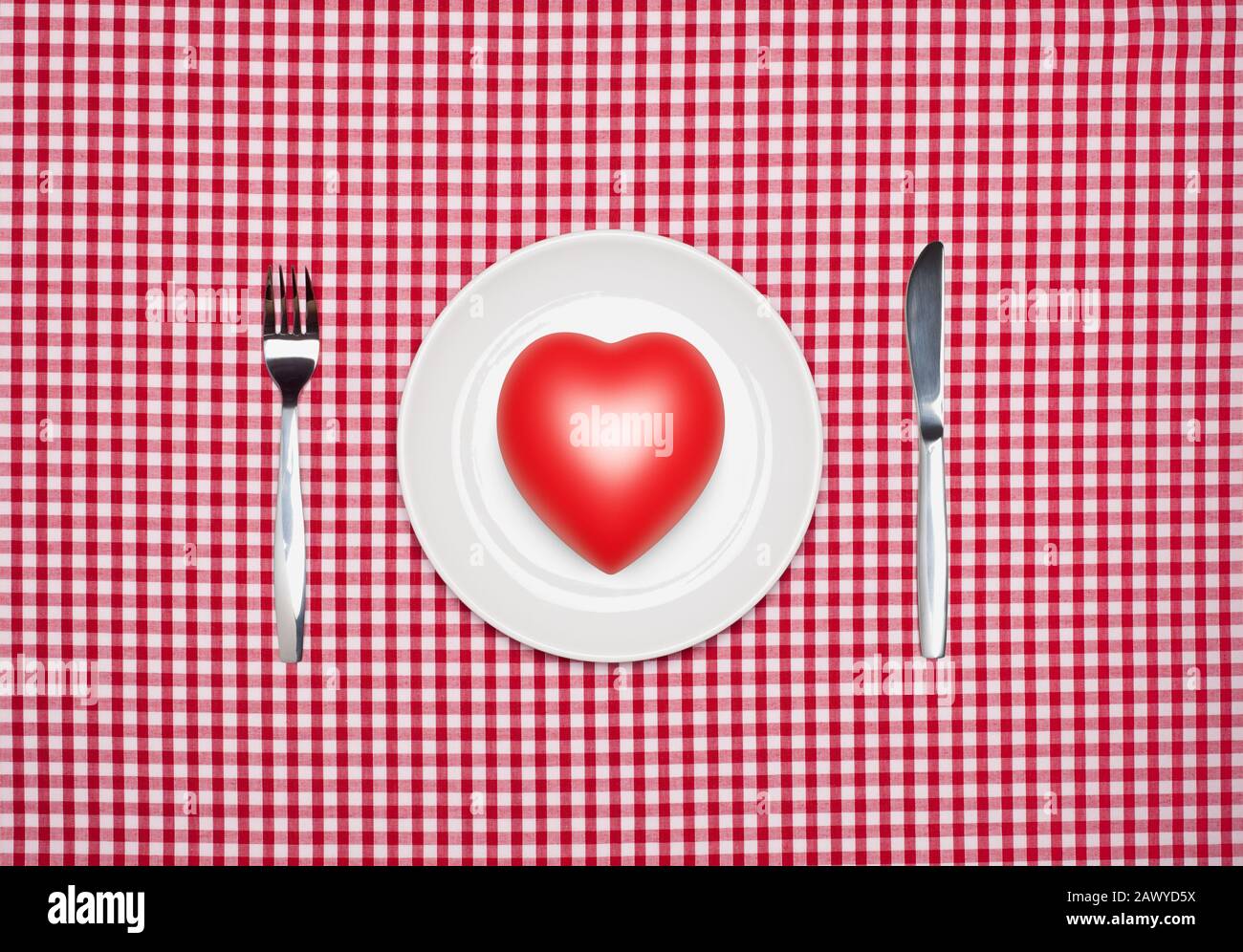 Healthy eating concept, a red heart on a round white plate with knife and fork from above on a red gingham tablecloth Stock Photo