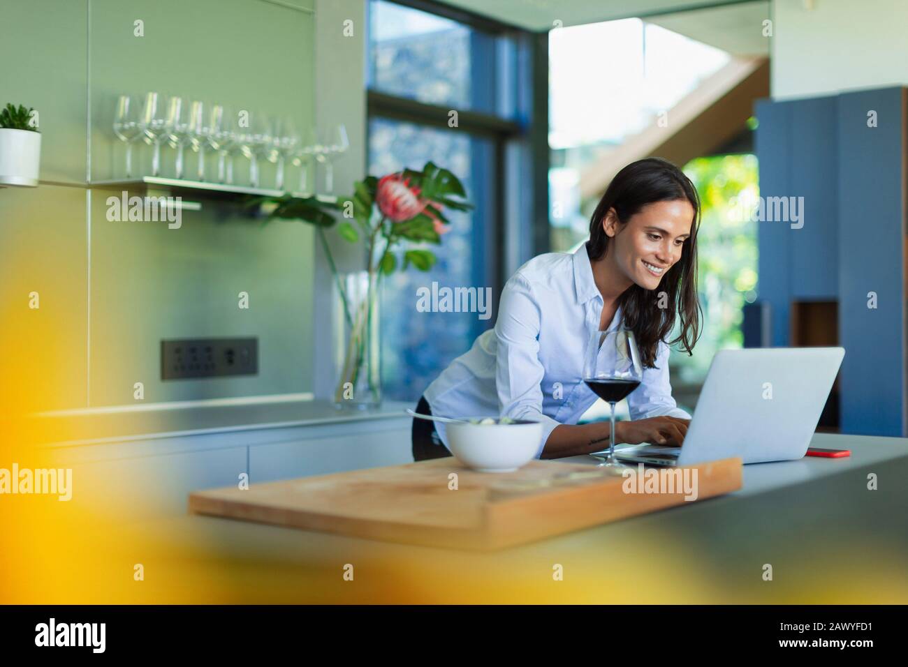 Smiling woman using laptop and drinking red wine in kitchen Stock Photo