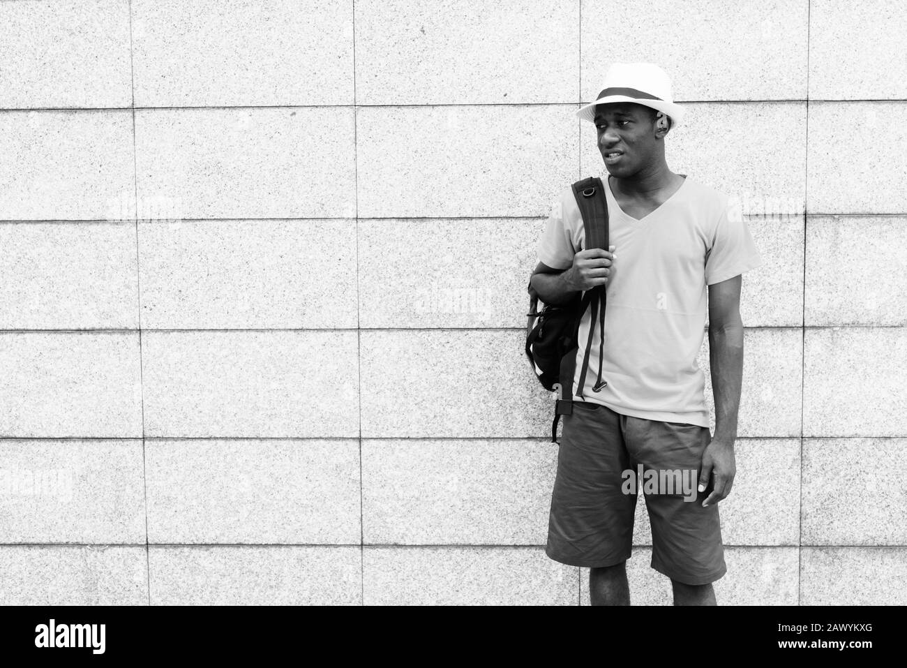 Young African tourist man standing and thinking while holding backpack against concrete block wall Stock Photo
