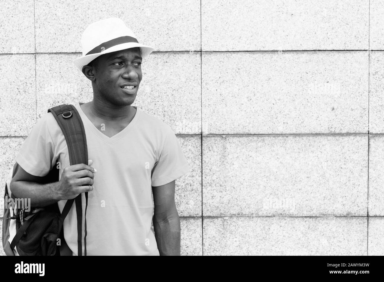 Young African tourist man holding backpack while thinking against concrete block wall Stock Photo