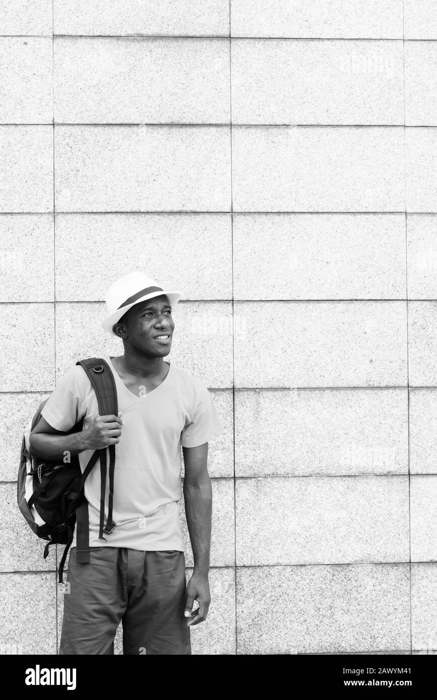 Happy young African tourist man thinking and holding backpack against concrete block wall Stock Photo