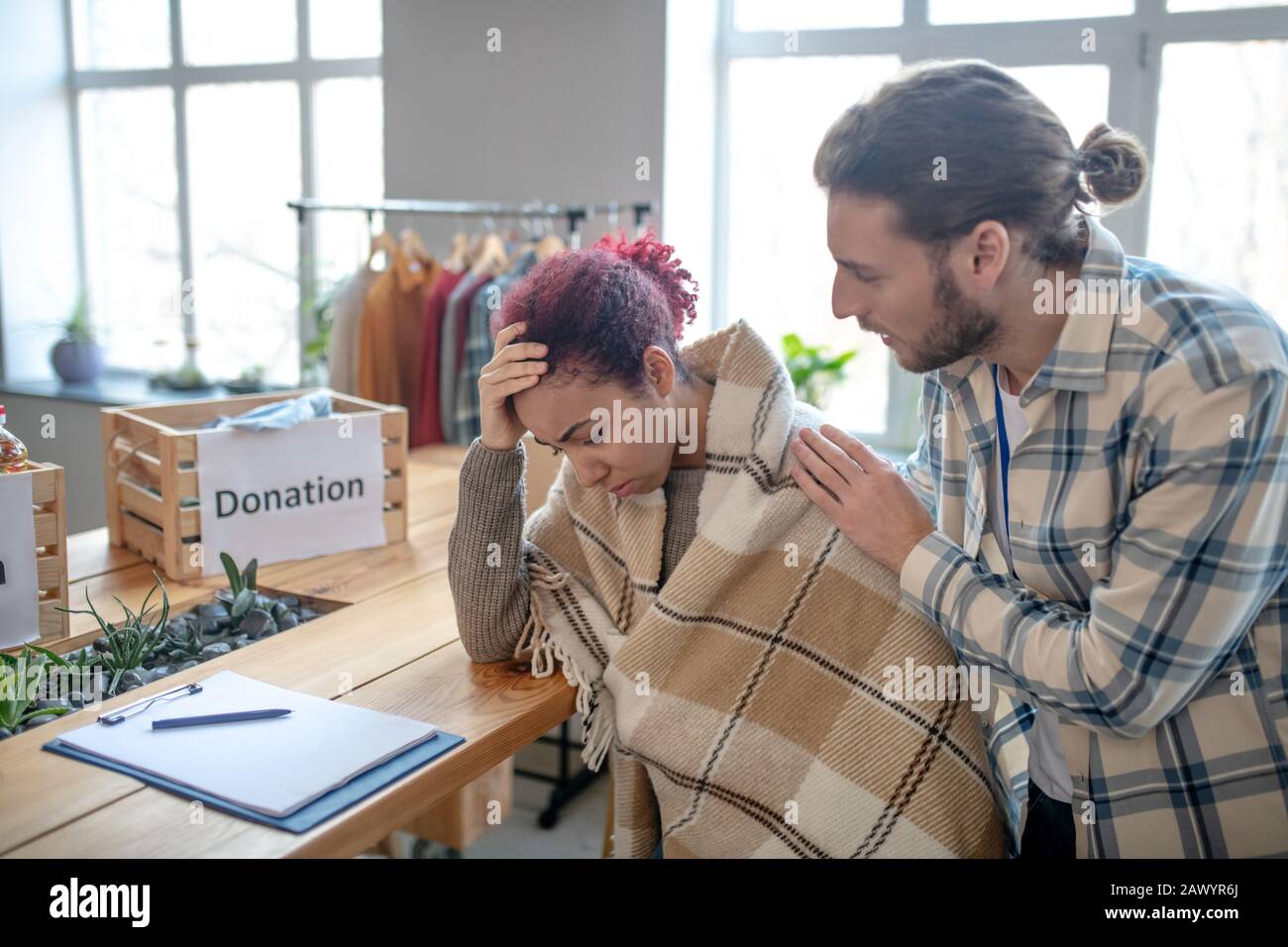 Young sad girl in plaid sitting tiredly at table, man hugging. Stock Photo