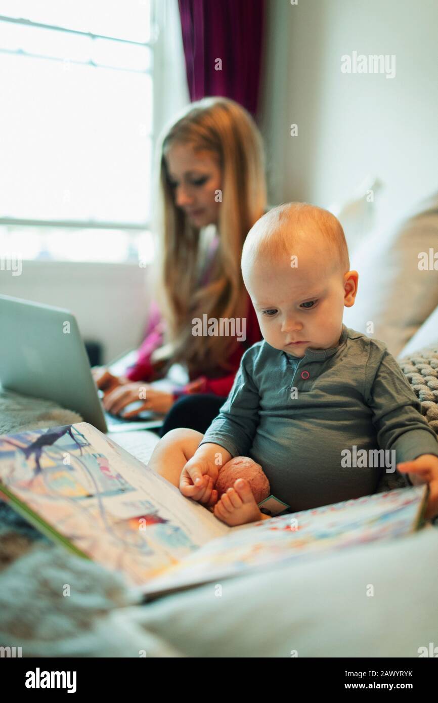 Cute innocent baby girl reading picture book on bed while mother works Stock Photo