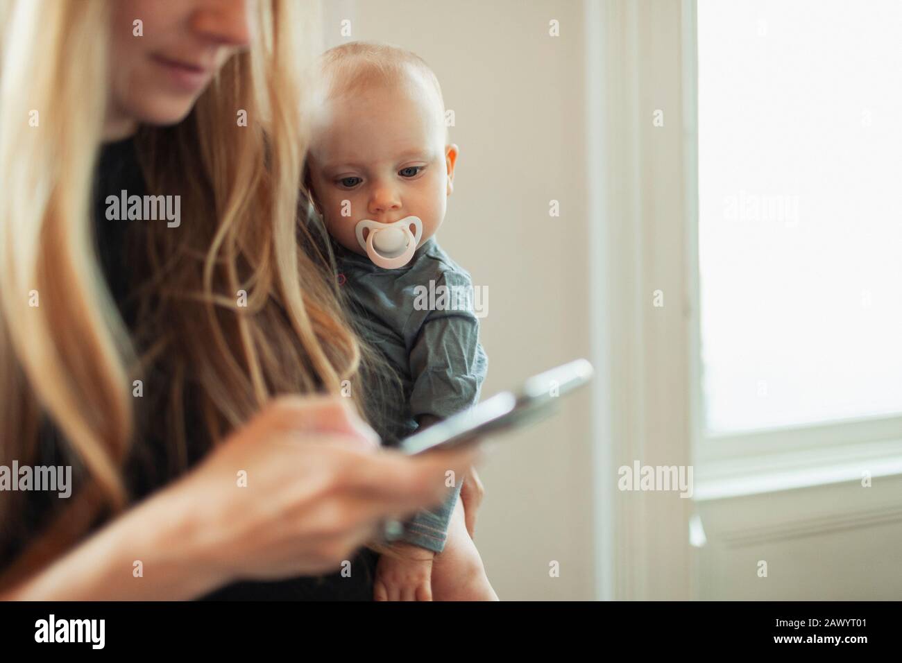 Curious baby girl with pacifier watching mother using smart phone Stock Photo