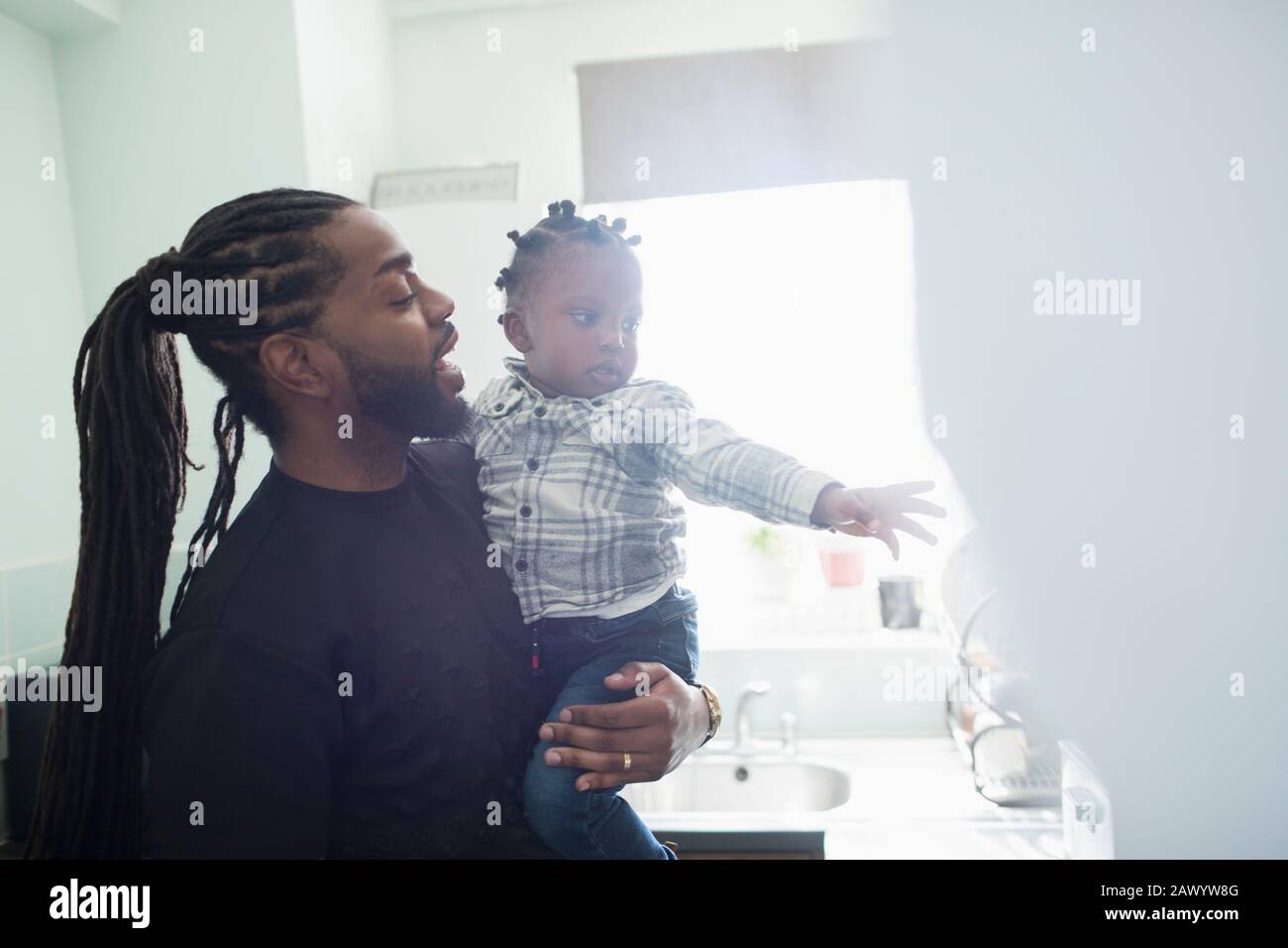 Father with long braids holding toddler son in sunny kitchen Stock Photo