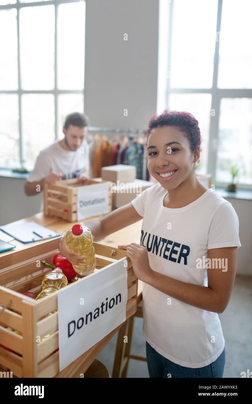 Young volunteer girl standing near food-filled donation box. Stock Photo