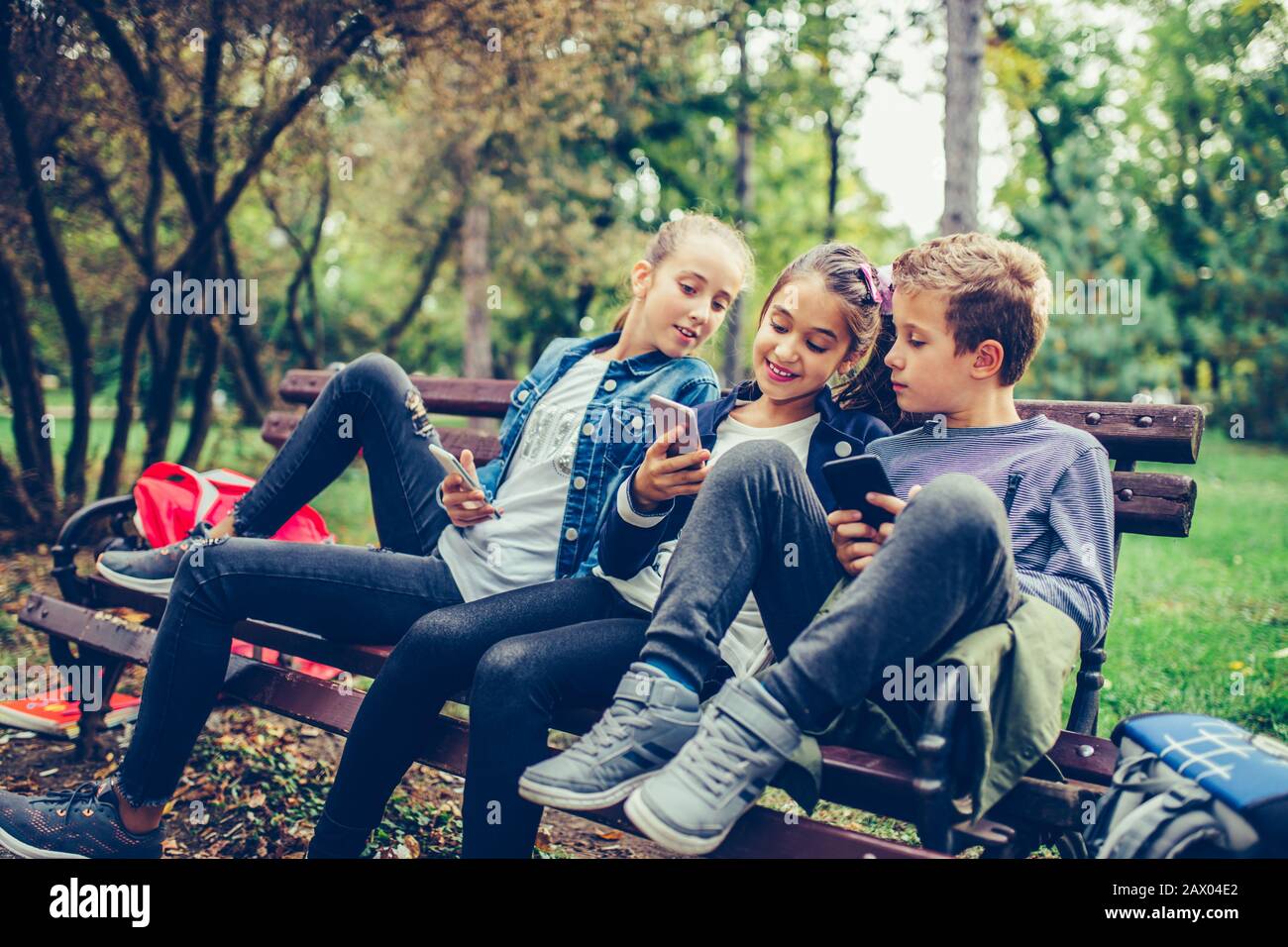 Group of friends playing video games on smart phone after school while sitting on the bench in the park. Stock Photo