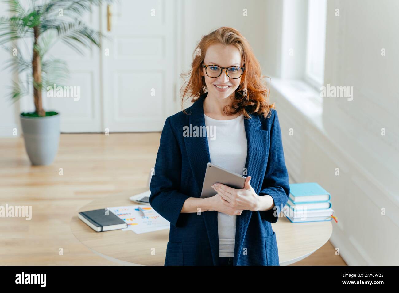 Horizontal shot of proud CEO checks email on digital touchpad, looks at camera with satisfied expression, wears spectacles, stands in modern office, e Stock Photo