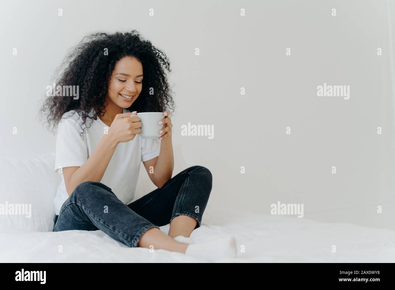 African American woman sips hot drink, poses in bed, enjoys good morning, dressed casually, spends weekend at home, blank space aside for your adverti Stock Photo