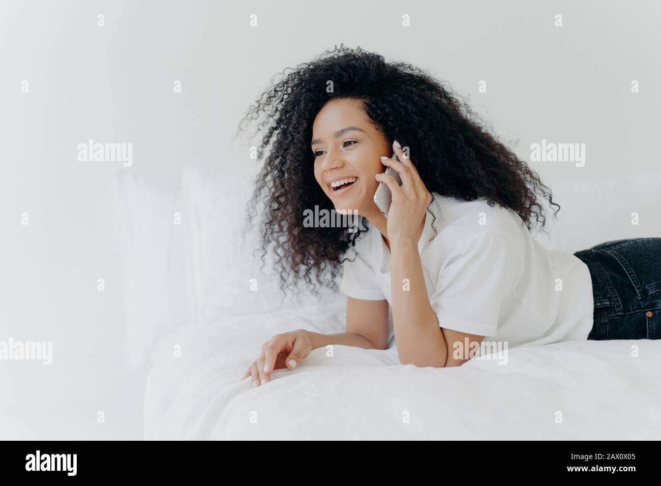 Charming young woman with crisp hair, speaks with boyfriend via smartphone, has rest in morning at bed, laughs at funny joke, dressed casually, being Stock Photo