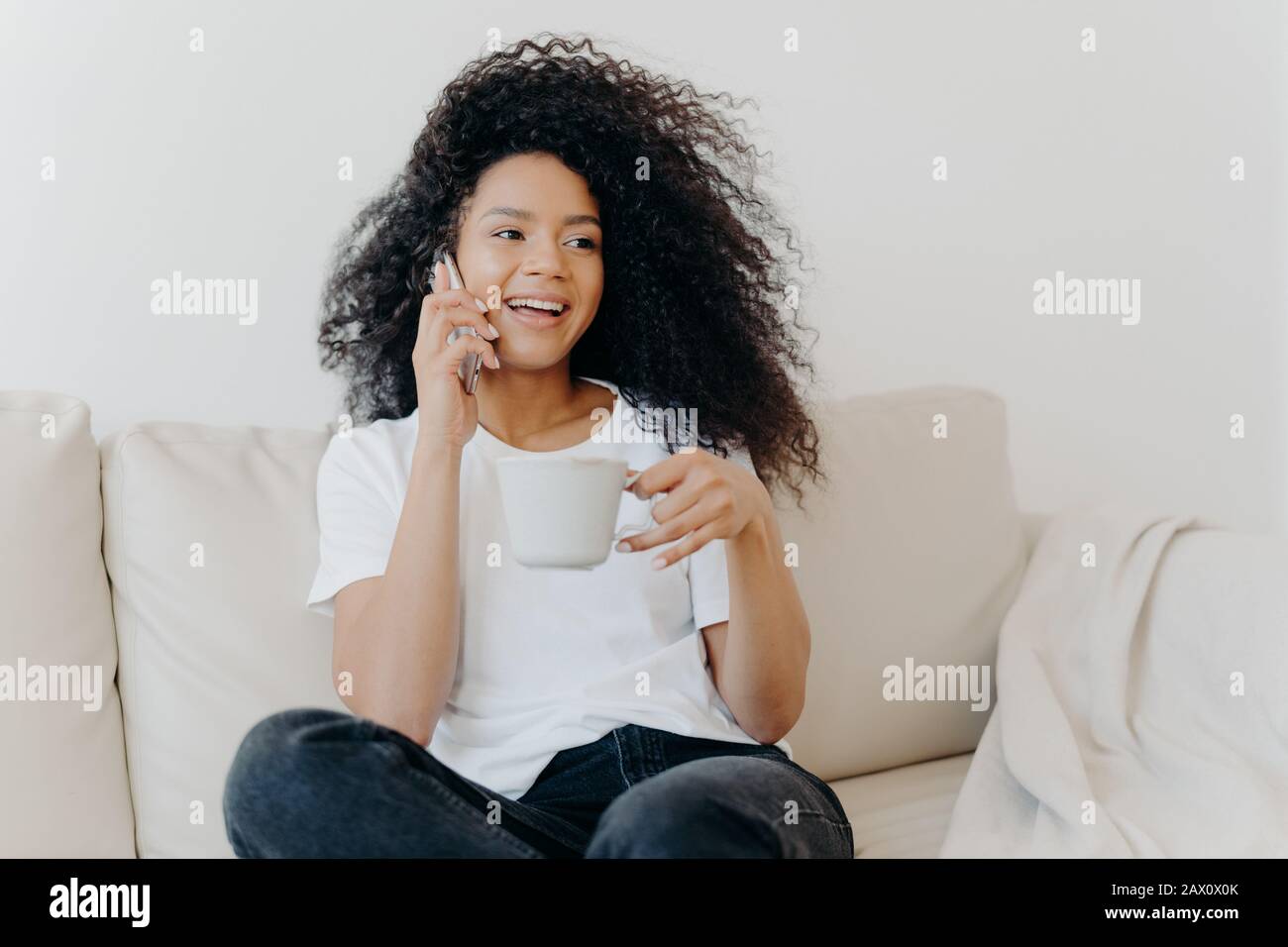 Smiling millennial girl with Afro hair, enjoys cellphone conversation with hot drink in cup, has healthy skin, dressed casually, sits on white sofa at Stock Photo
