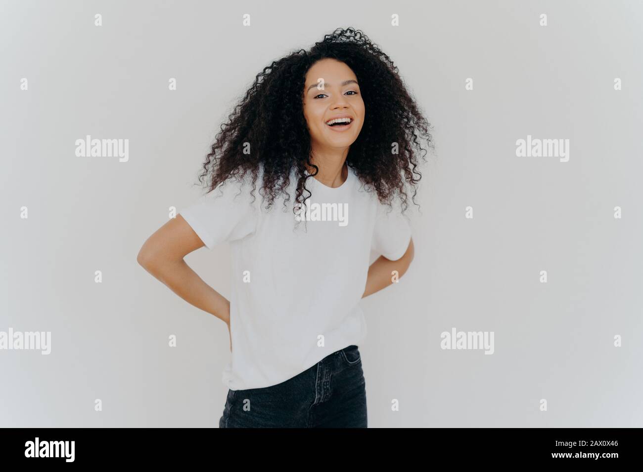 Portrait of cheerful millennial woman keeps hands behind back, dressed in casual wear, laughs from something fun, isolated over white studio backgroun Stock Photo
