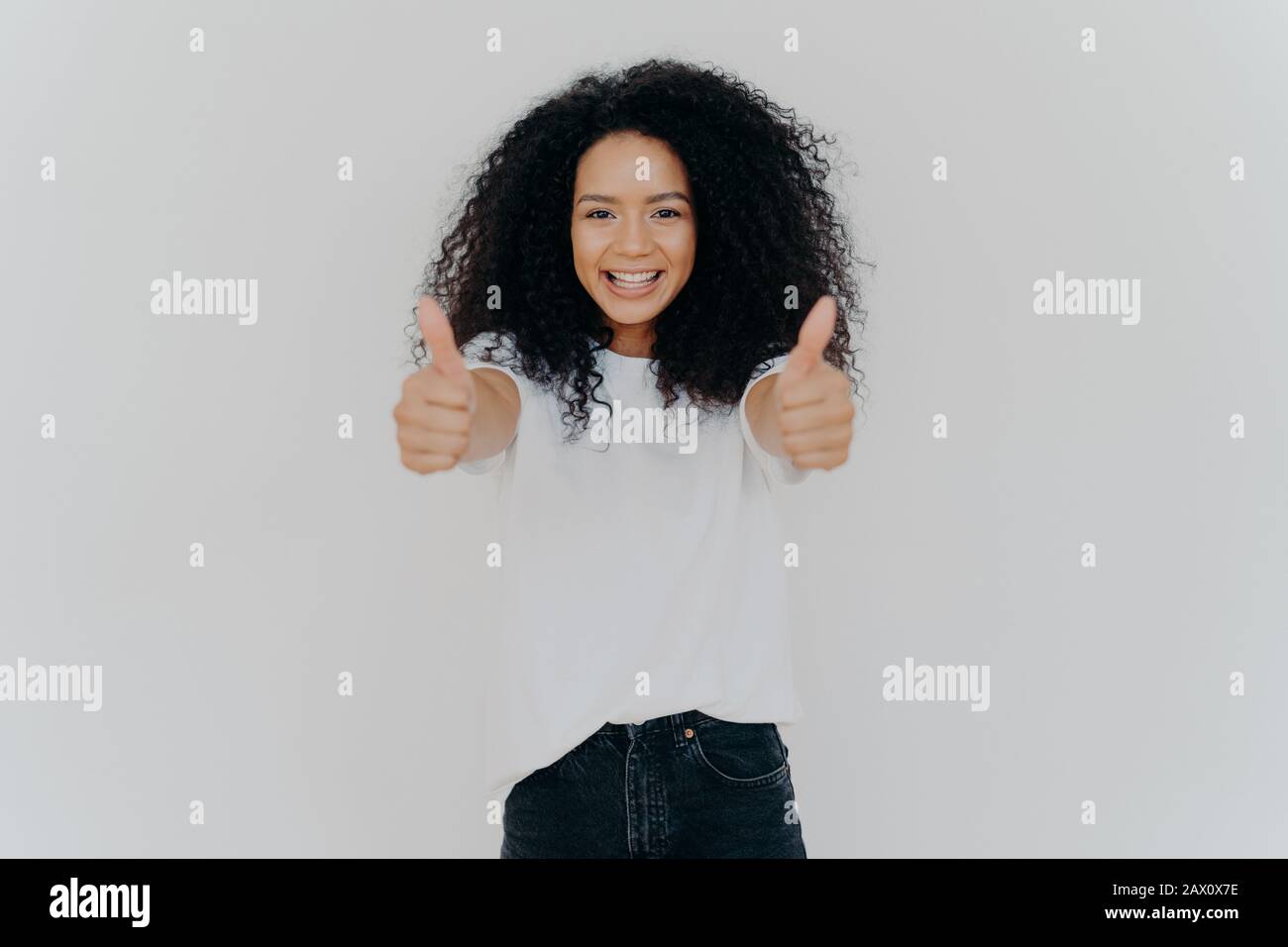 Happy young woman with Afro hairstyle, shows thumbs up, accepts or agrees awesome plan, gives her approval, smiles broadly, stands indoor, says excell Stock Photo