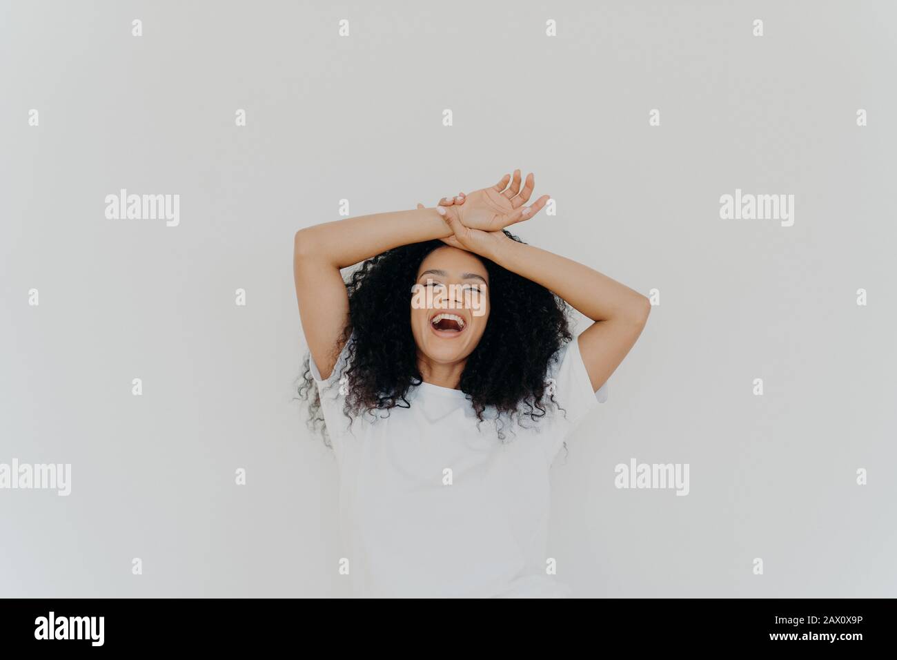 Overjoyed curly haired young lady keeps both hands on forehead, opens mouth and laughs happily, feels energetic, dressed in casual wear, poses against Stock Photo
